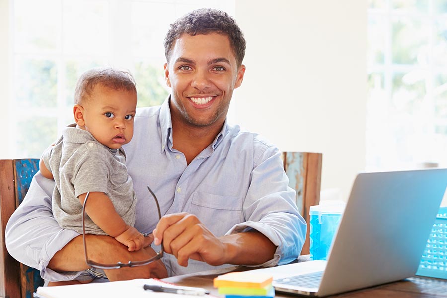 Client Center - Father Holding Baby Uses a Computer at the Kitchen Table
